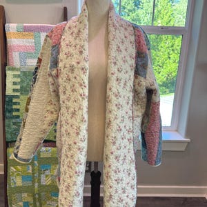 May include: A long-sleeved quilted jacket featuring a patchwork design with various floral and solid-colored fabric squares. The interior lining and wide lapels display a cream-colored fabric with a small pink and green floral pattern throughout.