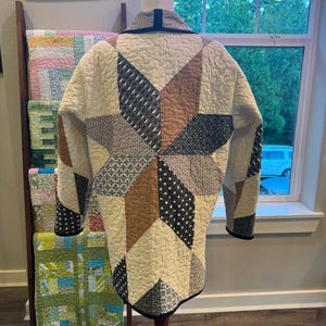 May include: A quilted jacket featuring a large star pattern on the back, constructed from various fabric panels in cream, tan, and dark blue with white geometric prints. The garment has a shawl collar, long sleeves, and black trim along the edges.