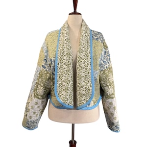 May include: A quilted jacket with a patchwork design in shades of green, blue, and cream. The jacket features a shawl collar with a light blue trim. The jacket is displayed on a mannequin.