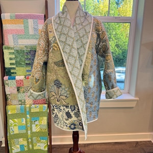 May include: A quilted jacket featuring a patchwork design with various floral and botanical patterns in shades of sage green, blue, and cream. The garment has a wide shawl collar, long sleeves with cuffs, and white piping trim along the edges.