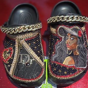 Op de afbeelding: Een paar zwarte klompen versierd met strass-steentjes, goudkleurige kettingbandjes en decoratieve bedels. De ene klomp heeft een ritsontwerp en een hartembleem, terwijl de andere een geborduurd portret toont van een persoon met een hoed en rode roos.