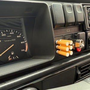 May include: Dashboard of a vintage car with a speedometer, gauges, and a cigarette holder with cigarettes. A Bic lighter is also visible. The dashboard is black, and the speedometer reads 33 mph.