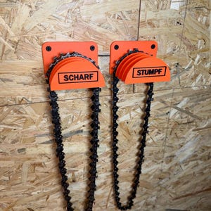 Könnte beinhalten: Zwei orangefarbene Kettensägenkettenhalter an einer Holzoberfläche befestigt. Jeder Halter hat ein halbkreisförmiges Design mit den Wörtern "SCHARF" und "STUMPF" in Schwarz. Die Ketten hängen herunter.