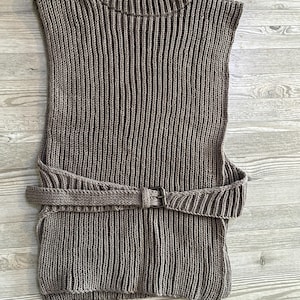 May include: A sleeveless, ribbed knit top in a muted brown color. The top features a high, ribbed neckline and a matching belt with a rectangular buckle. The vertical ribbing creates a textured appearance. The belt is wrapped around the waist.