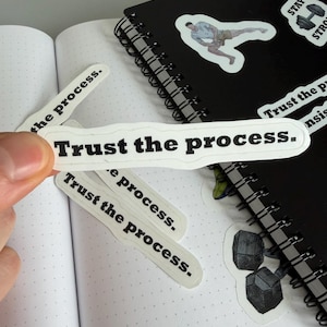 Può includere: Una mano tiene un adesivo bianco con la scritta "Trust the process" in nero. Diversi adesivi simili sono sparsi su una pagina di un quaderno a pois. Sullo sfondo, un quaderno nero a spirale con adesivi a tema fitness.