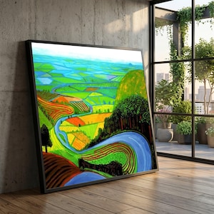 May include: A framed landscape painting depicting a vibrant, colorful valley with a winding blue river. The artwork features fields of green, yellow, and orange, with a blue sky and a forested area. The painting is displayed on a wooden floor.