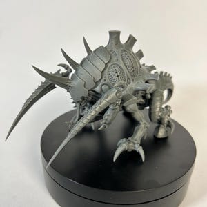 Op de afbeelding: Grijze plastic miniatuur van een Tyranid-wezen, met een gesegmenteerd lichaam, scherpe klauwen en meerdere stekelige uitsteeksels. Het model staat op een zwarte, cirkelvormige basis en toont zijn gedetailleerde ontwerp en dreigende uiterlijk.