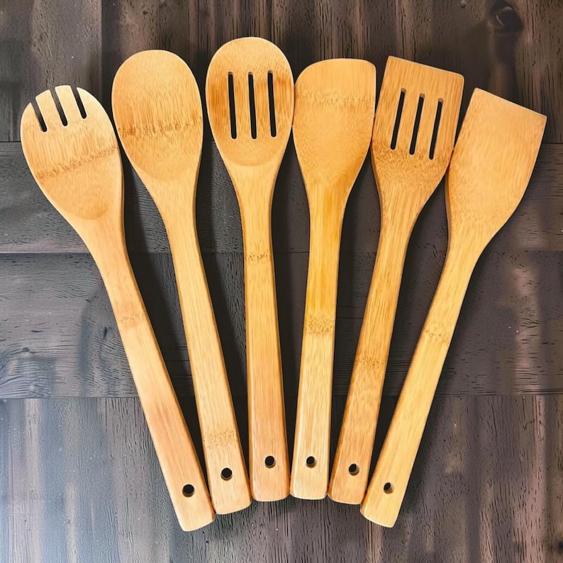 Puede incluir: Un juego de seis utensilios de cocina de bamb&uacute;, que incluyen una cuchara ranurada, una cuchara s&oacute;lida, una esp&aacute;tula y un tenedor para servir. Los utensilios de color marr&oacute;n claro est&aacute;n dispuestos sobre una superficie de madera oscura. Cada utensilio tiene un agujero en el mango.
