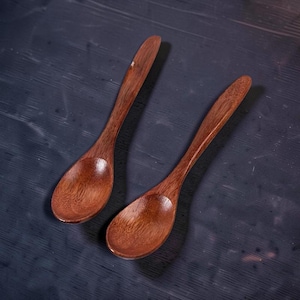 May include: Two wooden spoons with a deep brown colour. The spoons have an oval bowl and long, slender handles. The wood grain is visible, enhancing the natural look. Suitable for cooking and serving, each spoon measures approximately 20 cm in length.