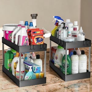 May include: Two black two-tier storage organisers with silver metal support poles. Each unit features a rectangular base and top shelf with integrated hooks, filled with various household cleaning supplies, plastic bottles, and detergent containers.