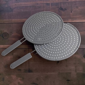 May include: Two gray splatter screens with handles. Each screen is round with numerous small holes. The handles are also gray. The screens are on a dark wood surface.