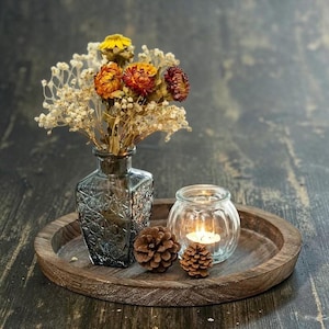 May include: A round wooden tray holding a square, textured gray glass vase filled with dried yellow, orange, and burgundy flowers and white baby's breath. Next to the vase are two brown pinecones and a small, clear, ribbed glass candle holder.