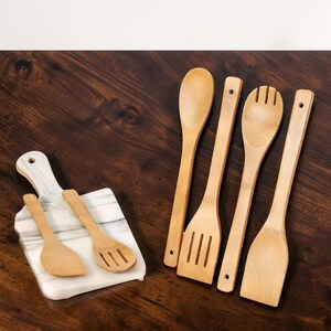 Puede incluir: Un conjunto de utensilios de cocina de bamb&uacute;, que incluye una cuchara, una cuchara ranurada, una esp&aacute;tula y una cuchara de servir, junto con una peque&ntilde;a tabla de cortar de m&aacute;rmol con dos utensilios de bamb&uacute;. Los utensilios son de color marr&oacute;n claro.