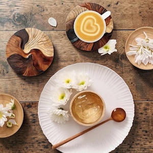 May include: A white fluted plate holds a small wooden bowl filled with clear liquid and a long wooden spoon. Nearby are a wooden coaster with a heart-patterned latte, a carved wooden disc with swirling grain, and small bowls containing white flowers.