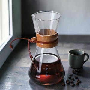 May include: A clear glass coffee maker with a wooden collar and leather strap, filled with brewed coffee. A small green mug and coffee beans are next to the coffee maker. The coffee maker has measurement markings.