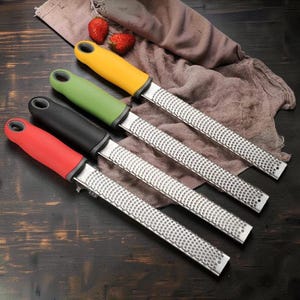 Könnte beinhalten: Vier Küchenreiben mit langen, rechteckigen Metallklingen und kleinen, gleichmäßigen Perforationen. Jede Reibe hat einen farbigen, ergonomischen Griff in Gelb, Grün, Schwarz oder Rot sowie eine kleine, runde Öffnung am Ende zum Aufhängen.