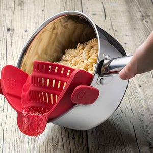 May include: A stainless steel saucepan containing cooked rotini pasta, fitted with a red silicone clip-on strainer attached to the rim. The strainer features vertical slots and a pouring spout, allowing water to drain while keeping the pasta inside.