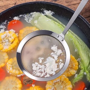 May include: A stainless steel skimmer spoon with a fine mesh circular head, positioned over a dark speckled cooking pot filled with broth, sliced corn on the cob, green leafy vegetables, and orange carrot slices, capturing white food foam.