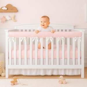 May include: A white crib with a pink polka dot bumper and matching bedding. A baby in a white onesie is standing in the crib. The room is decorated with a rainbow art print and a wooden cloud mobile.