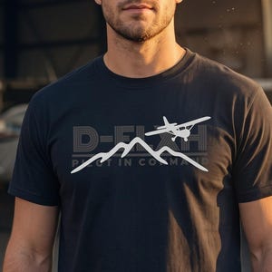 Peut inclure: Un t-shirt noir à manches courtes avec un imprimé graphique blanc sur la poitrine. Le motif représente un petit avion volant au-dessus d'une silhouette de montagne avec le texte D-FLY et PILOT IN COMMAND en police sans empattement.