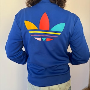Puede incluir: Chaqueta deportiva azul real con cierre de cremallera completa y cuello alzado. La parte trasera presenta un gran logotipo colorido con detalles en naranja, rojo, amarillo, rosa y turquesa. La chaqueta es de estilo deportivo.