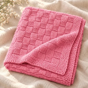 Peut inclure: Une couverture tricotée rose vif présentant un motif en damier texturé. Le textile de forme carrée est plié soigneusement, mettant en valeur un point de tricot uniforme sur toute la surface, y compris sur les bords du tissu.