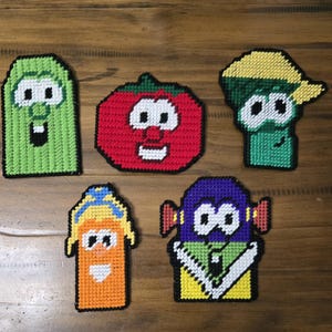Puede incluir: Cinco figuras coloridas hechas a mano de lona plástica de verduras de dibujos animados. Las figuras incluyen un pepino verde, un tomate rojo, un brócoli verde, un personaje naranja y un personaje morado. Cada figura tiene una cara sonriente.