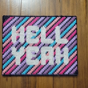 May include: A colorful cross-stitch piece with a striped background and the words "Hell Yeah" in white.