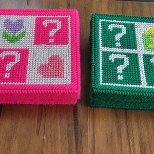 Könnte beinhalten: Zwei kleine Boxen mit einem Gittermuster auf der Oberseite. Die Box auf der linken Seite ist pink mit einer Blume, einem Herzen und Fragezeichen. Die Box auf der rechten Seite ist grün mit einer Ampel, einem Block und Fragezeichen.