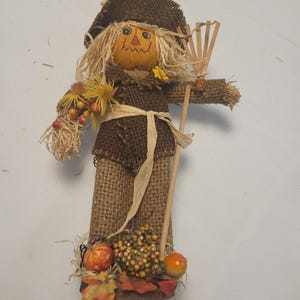 May include: A scarecrow figurine crafted from brown burlap fabric, featuring a round orange face with painted features, a wide-brimmed hat, and straw hair. It holds a wooden rake and is decorated with artificial yellow flowers, berries, and pumpkins.