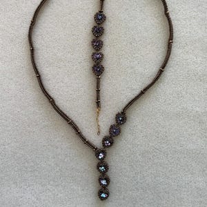 Op de afbeelding: Een sieradenset bestaande uit een ketting en een armband. De ketting en armband zijn gemaakt van bruine kralen en hebben clusters van paarse en iriserende kralen. De ketting heeft een Y-vormig ontwerp en de armband heeft een eenvoudige ketting.