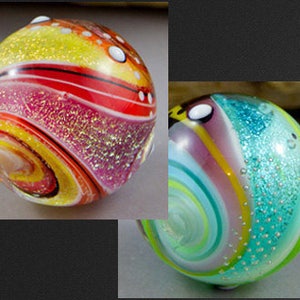 Può includere: Due biglie di vetro colorate con motivi a spirale e accenti di glitter. Una biglia è rossa, gialla e bianca, mentre l'altra è blu, verde e viola.