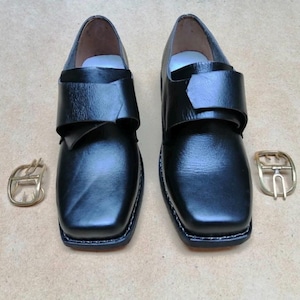 Pode incluir: Um par de sapatos de couro preto com biqueira quadrada e uma tira larga na parte superior. Cada sapato apresenta uma sola grossa com costuras visíveis ao longo da borda. Duas fivelas de metal cor de latão estão colocadas ao lado dos sapatos.