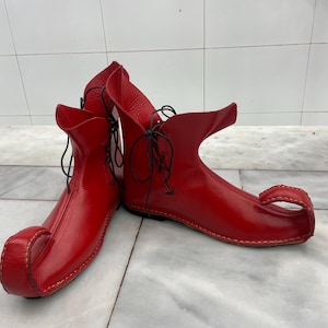 Op de afbeelding: Een paar felrode leren laarzen met een uniek, puntig teenontwerp. De laarzen hebben een hoge, open manchet en zijn geregen met zwarte veters. De stiksels zijn zichtbaar en de laarzen lijken handgemaakt.