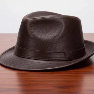 May include: A dark brown fedora hat with a smooth, textured finish. The hat features a structured crown with a central crease and a matching dark brown band encircling the base. The brim is slightly curved with visible stitching along the edge.