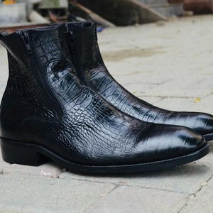 Pode incluir: Um par de botas de tornozelo pretas de couro com um padrão texturizado semelhante ao de um crocodilo. As botas têm zíperes laterais e um salto baixo. O calçado é exibido em uma superfície de pedra.