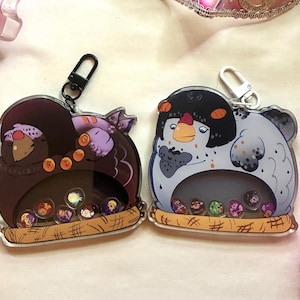 May include: Two acrylic keychains featuring cartoon bird designs. One is dark purple with orange accents, the other is light blue with black accents. Both have a clear center with small, colorful character images and a brown nest base.