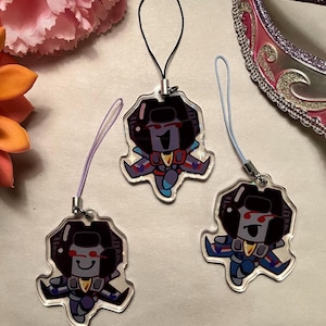 May include: Three clear acrylic charms featuring a cartoon character design with a black, red, blue, and yellow color scheme. Each charm has a different colored lanyard attached. The charms are on a white surface.
