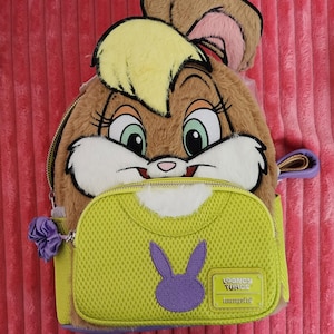 May include: A brown and yellow Lola Bunny backpack with a green pocket featuring a purple bunny silhouette. The backpack has a zipper closure and a small purple bow. The Looney Tunes Loungefly logo is visible.