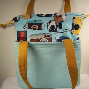May include: A light blue tote bag with brown straps. The bag has a fabric lining with a pattern of vintage cameras and a light blue background.