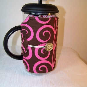 May include: A French press coffee maker with a black handle and lid. The glass carafe is wrapped in a brown fabric with a pink swirl pattern, secured with a gold button. The coffee maker sits on a white surface.