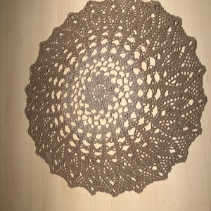 Könnte beinhalten: Handgefertigte, taupefarbene Häkelspitze. Die kreisförmige Spitze weist ein kompliziertes Lochmuster mit einem gewellten Rand auf. Die Spitze ist aus weichem, strukturiertem Garn gefertigt und wird auf einer hellen Holzoberfläche präsentiert.
