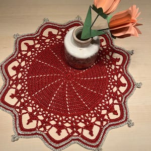 Może przedstawiać: Czerwona szydełkowa serwetka ze srebrną obwódką i falistym brzegiem. Mały biały wazon z brązową obwódką trzyma dwa papierowe kwiaty. Kwiaty są w kolorze brzoskwiniowym z zielonymi papierowymi liśćmi.