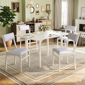 May include: A white dining table with four chairs. The chairs have light gray upholstered seats and backs. The table is set with plates, silverware, and a vase of flowers. The room has a neutral color palette with a patterned rug.