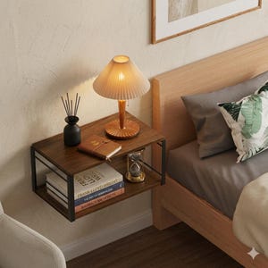 May include: A floating nightstand with a dark wood finish and black metal frame. It features a lamp, books, a journal, a black vase, and an hourglass. The lamp has a pleated shade and a wooden base. The nightstand is next to a bed with gray bedding.