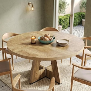 May include: A large, round wooden dining table with a light brown finish. The table is surrounded by wooden chairs with beige cushions. A teal bowl with bread, plates, and a candle are on the table. The table sits on a beige rug.