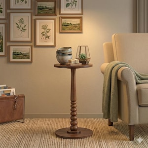 May include: A small, dark brown wooden side table with a round top and base. The table features a decorative, beaded column. On top are decorative bowls, soaps, and a geometric terrarium with a small plant. The table is next to a beige armchair.