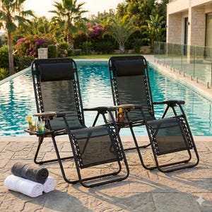 May include: Two black zero-gravity lounge chairs with attached side tables, positioned near a pool. Each chair has a black headrest and armrests. Rolled white, gray, and white towels are on the ground. Drinks and sunscreen are on the side tables.
