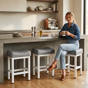 May include: Three gray upholstered bar stools with white wooden frames are arranged in front of a concrete countertop. The stools have a curved seat design. A woman is sitting on one of the stools.