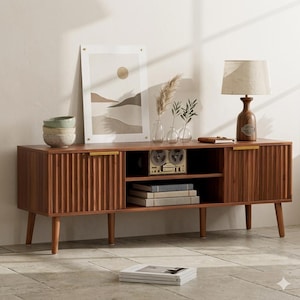 May include: A mid-century modern style wooden TV stand with a warm brown finish. The stand features fluted door panels, gold-toned handles, and tapered legs. It has open shelving and a lamp with a beige shade. Decorative items include artwork, vases, and books.